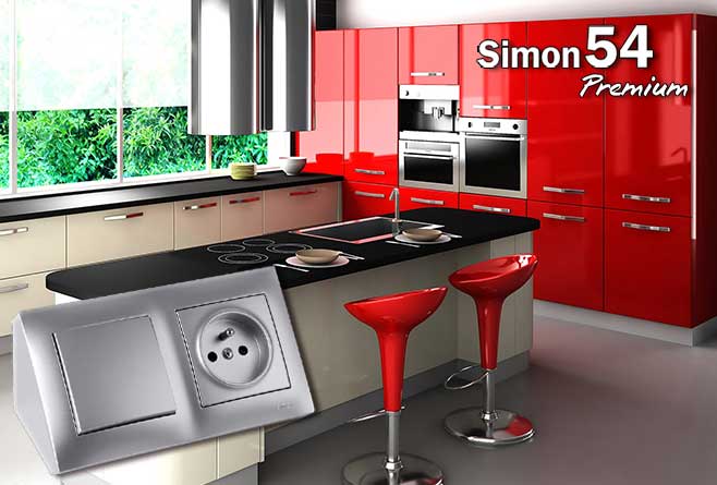 Nowoczesna kuchnia z czerwonymi frontami i wyspą kuchenną, na pierwszym planie srebrny osprzęt Simon 54 Premium z gniazdkiem i łącznikiem.